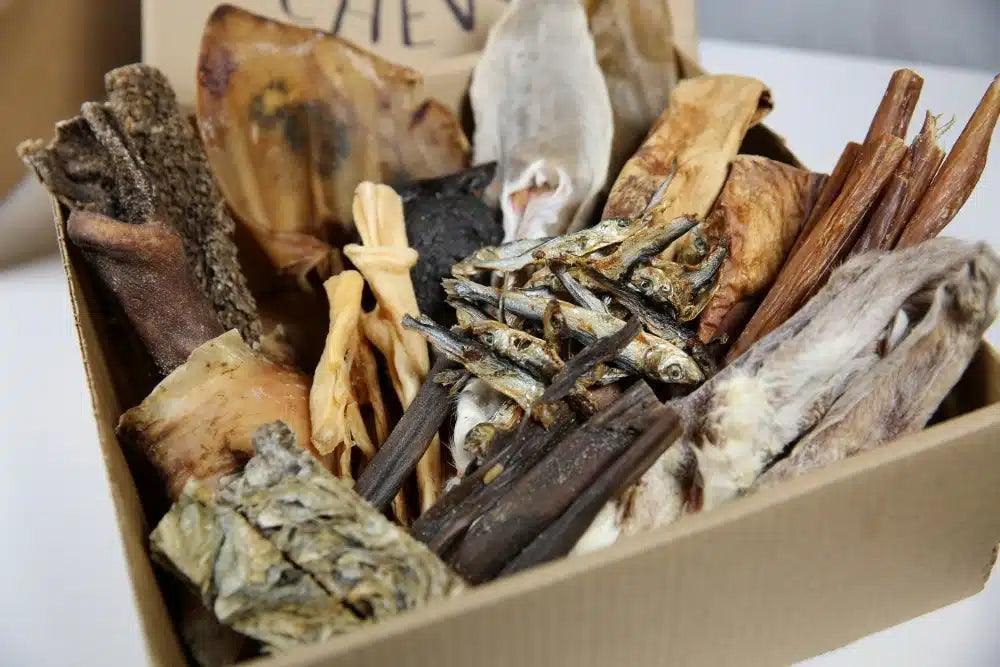 Box of assorted natural dog treats including dried fish, bones, and chews in a cardboard container